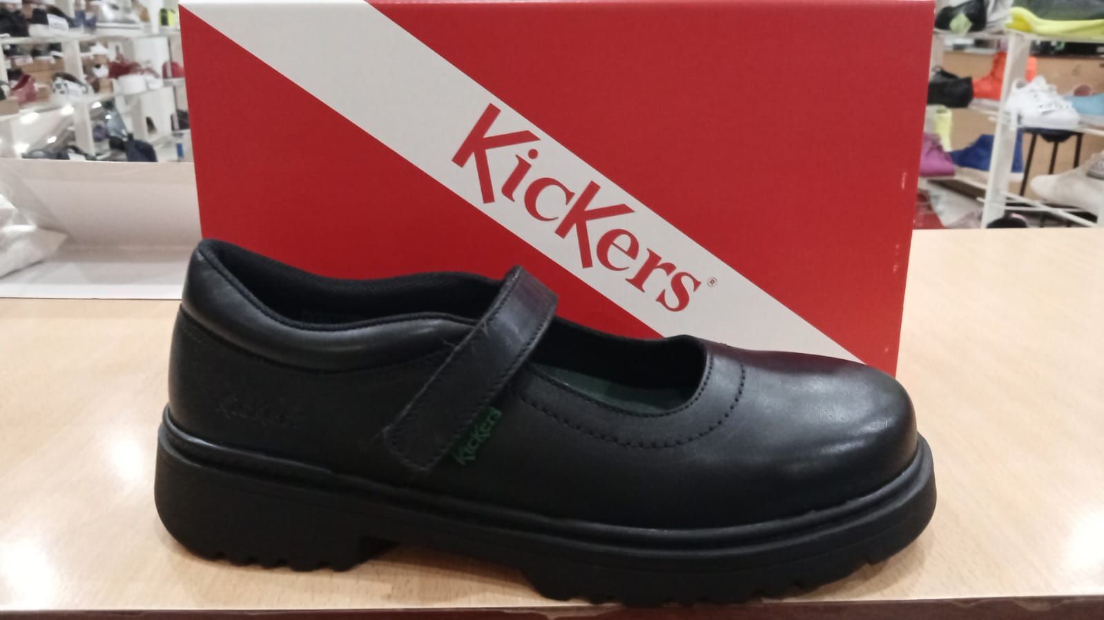 Zapato escolar Kickers negro delante de una caja Kickers roja y blanca sobre una mesa de madera.