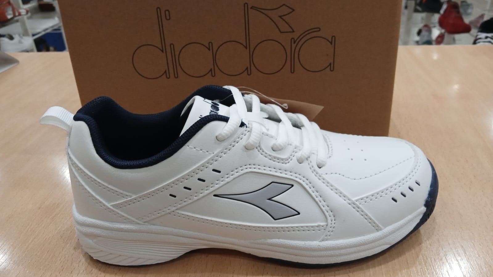 Zapatilla Diadora blanca con detalles en azul marino, delante de su caja de cartón.