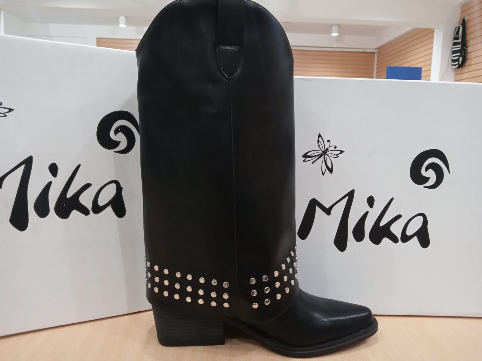 Bota vaquera negra hasta la rodilla con detalle de tachuelas en el tobillo, expuesta frente a un letrero de Mika.