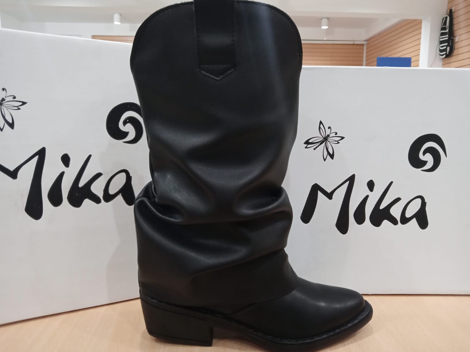 Bota negra hasta la rodilla expuesta frente a un fondo con la marca Mika.