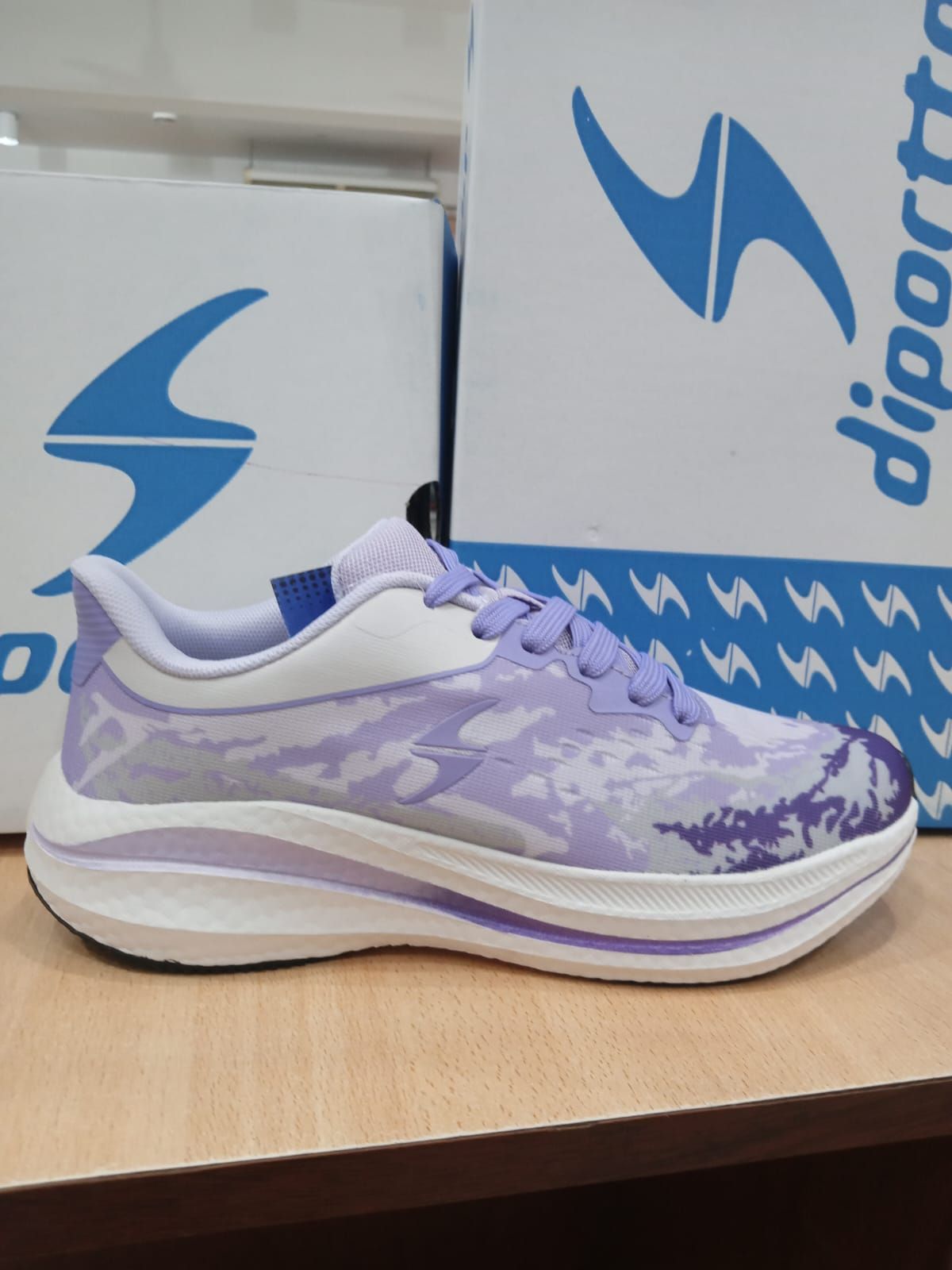 Zapatillas deportivas moradas y blancas expuestas sobre una superficie de madera con cajas de marca al fondo.