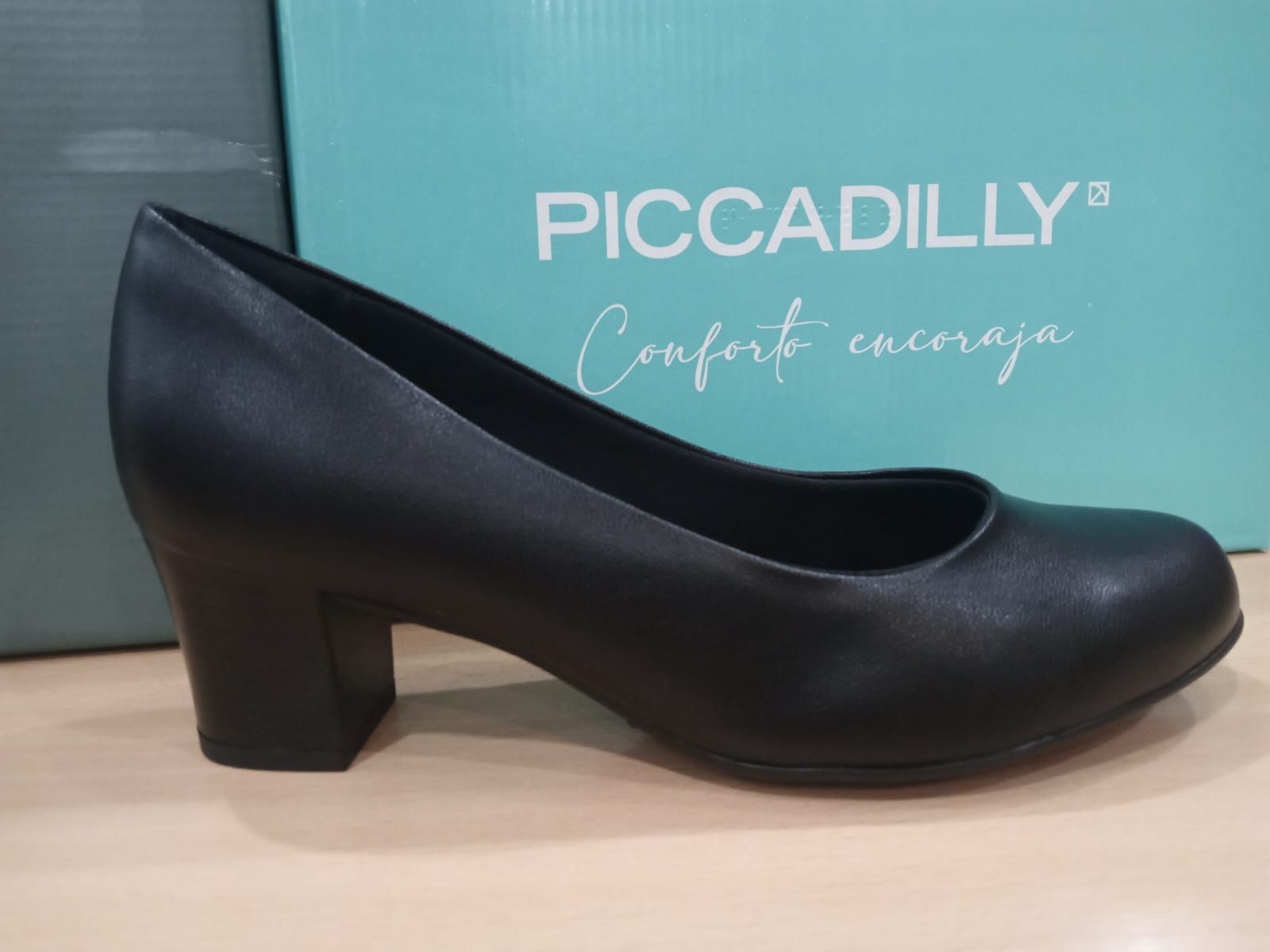 Un zapato de tacón negro sobre un estante con una caja de zapatos Piccadilly al fondo.