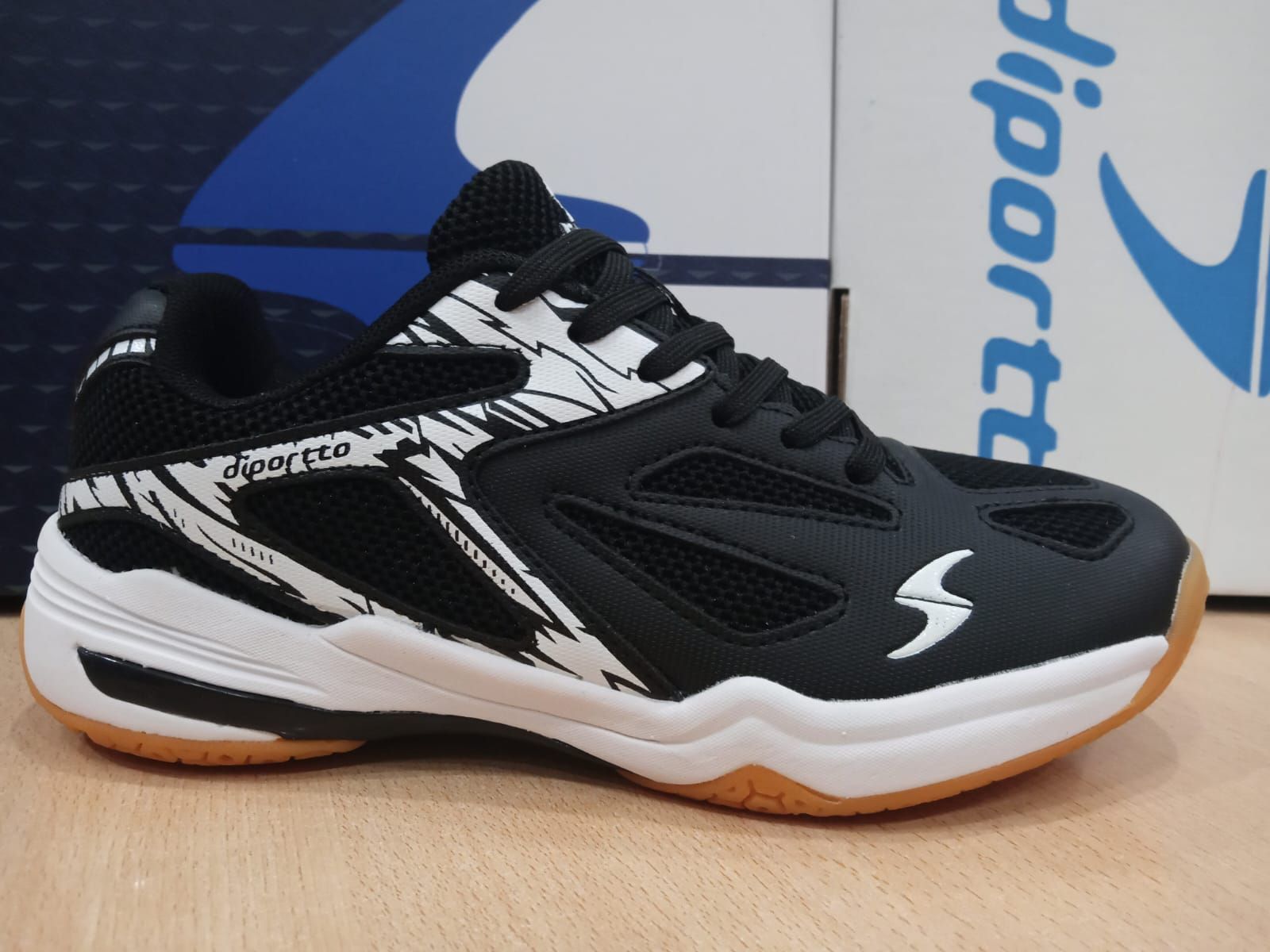 Zapatillas deportivas blancas y negras sobre una superficie de madera, con suela naranja y caja azul detrás.