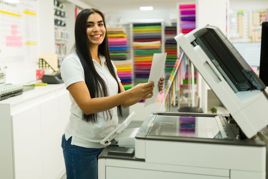 A smiling person in a white polo shirt places a document onto the glass of a large office photocopier in a print shop.