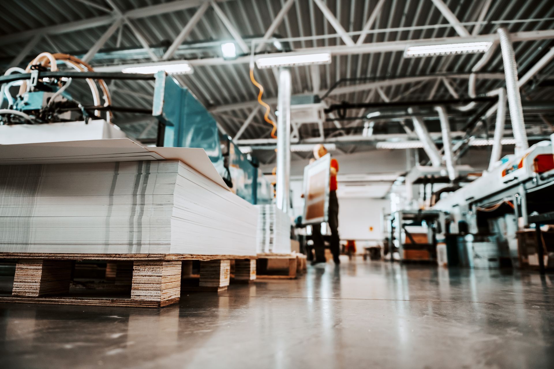 A worker moves material in a printing factory with a large stack of white paper on a pallet in the foreground.