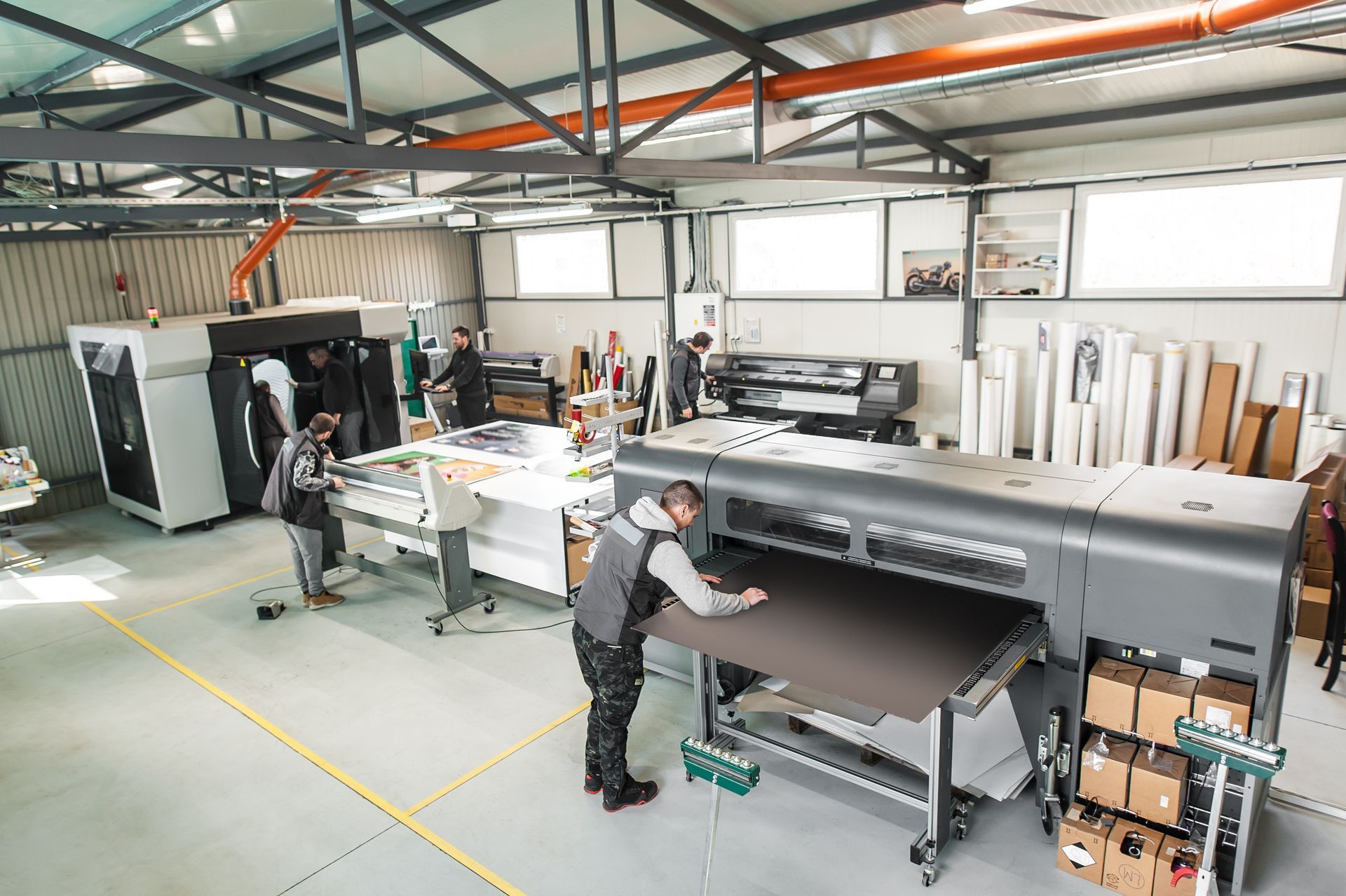 Workers operate industrial printing machinery inside a brightly lit warehouse studio with materials stored in the back.