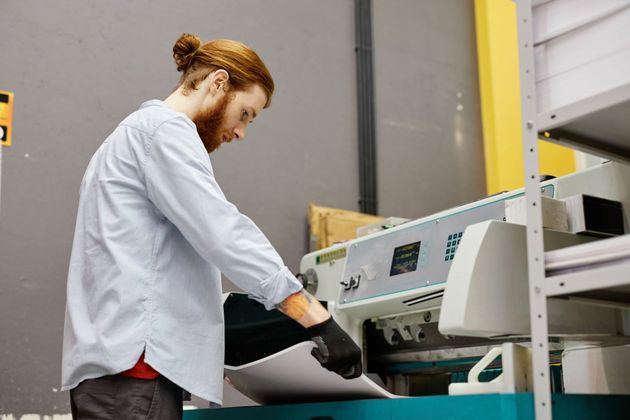 A person wearing a light blue shirt and black gloves feeds a sheet of white paper into a printing machine.