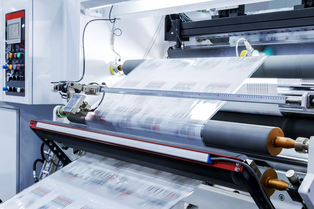 An industrial machine processing a continuous roll of printed plastic film through rollers in a factory setting.