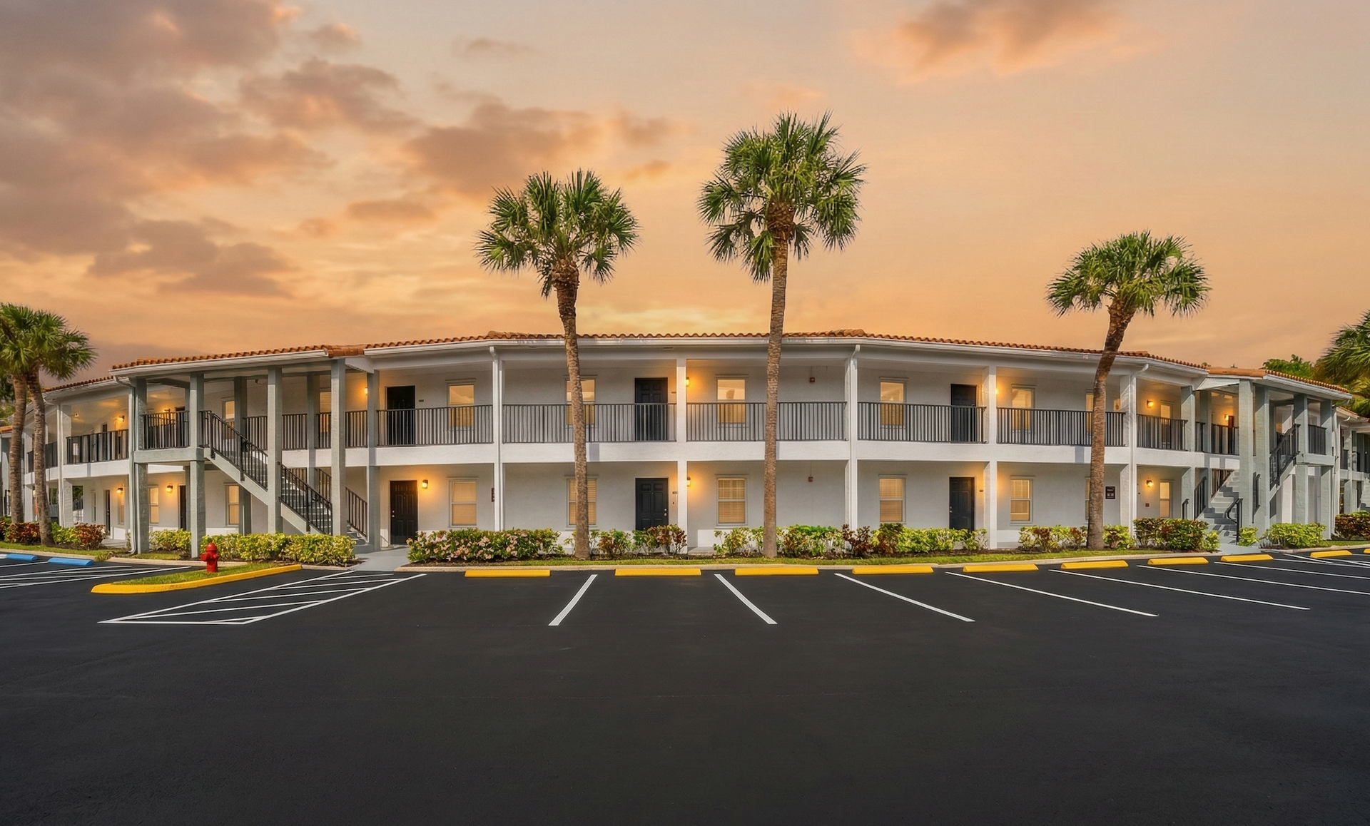 A two-story, white apartment building with a palm-lined parking lot under a sunset sky.
