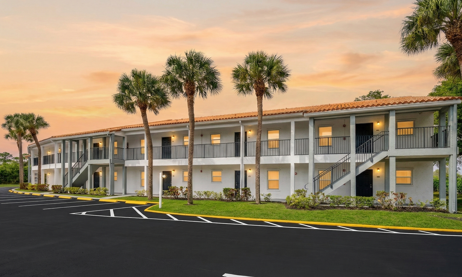 A two-story white apartment building with balconies and palm trees under a sunset sky.