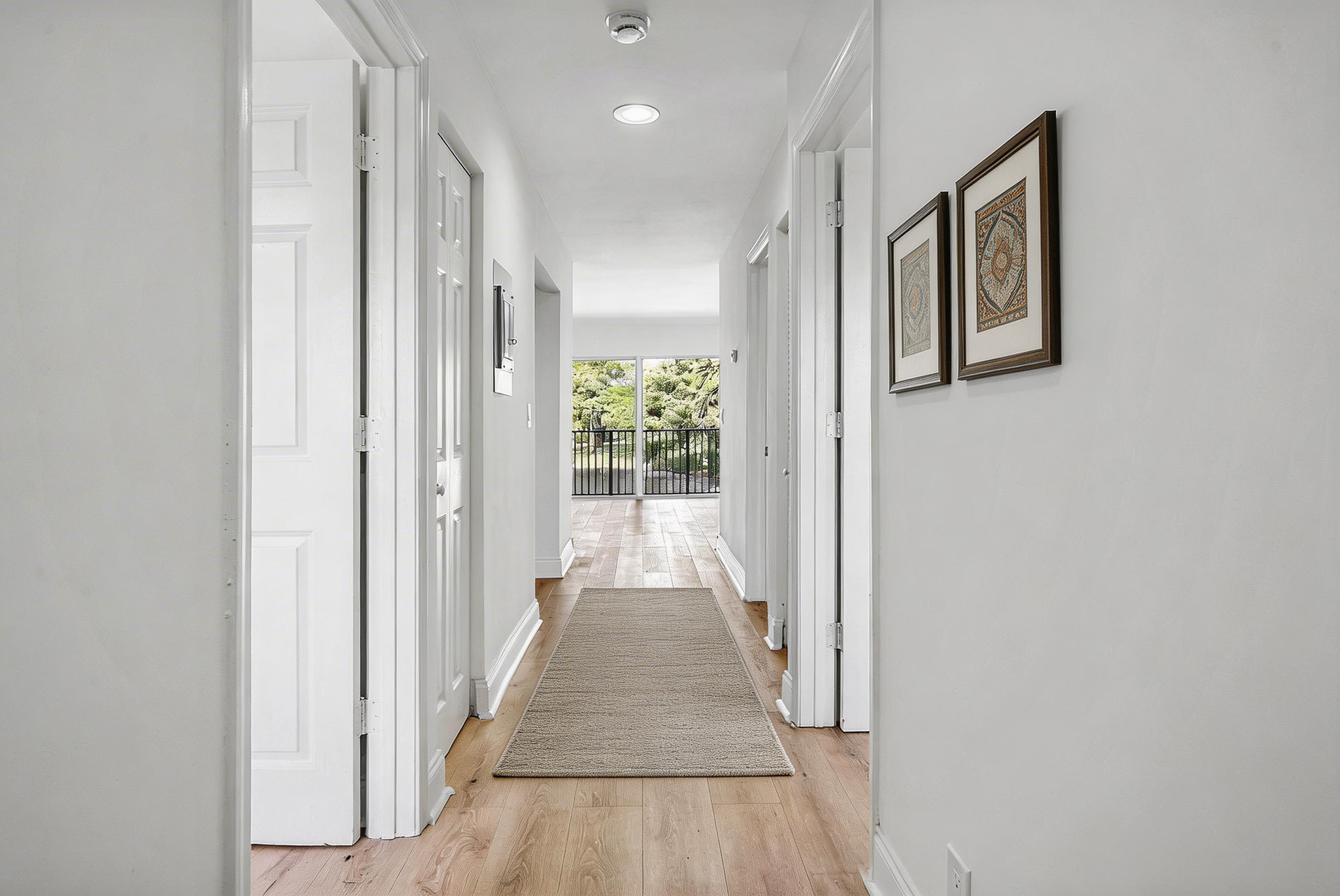 A bright hallway with white walls and wood-look flooring, leading to a balcony view through an open doorway at the end.