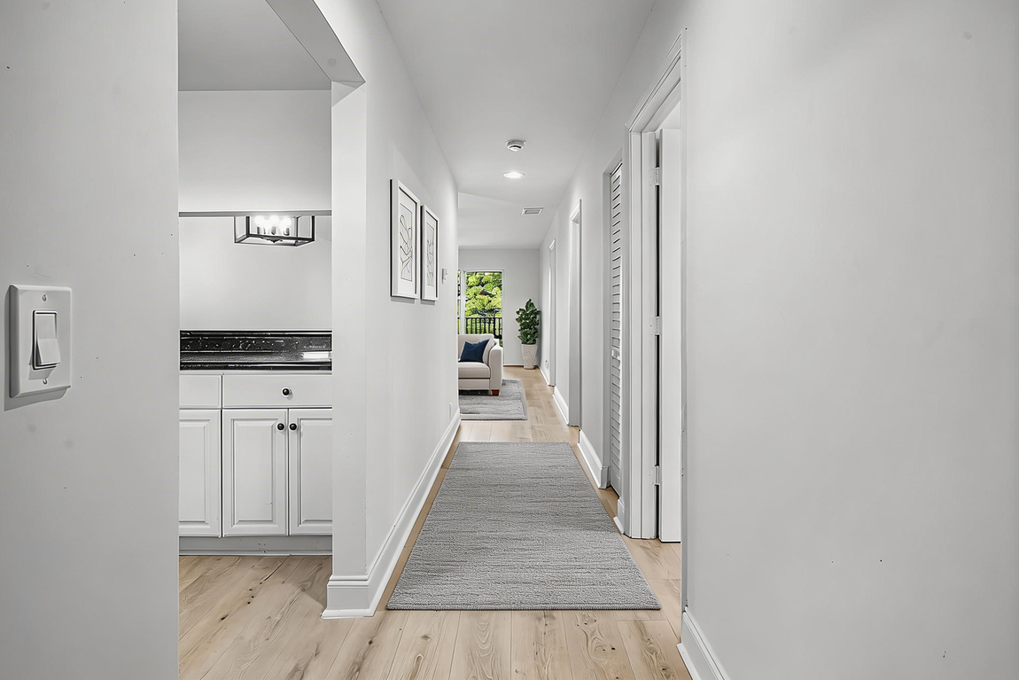 A bright, white hallway with light wood floors, a grey patterned runner rug, and an open doorway leading to a vanity.