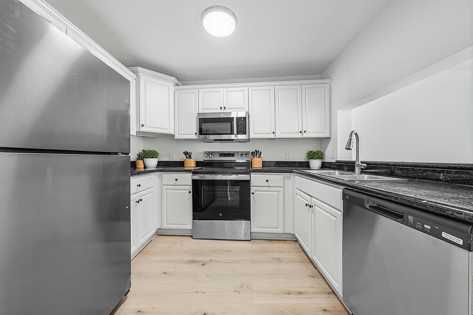 A bright kitchen with white cabinets, stainless steel appliances, dark countertops, and light-colored wood flooring.