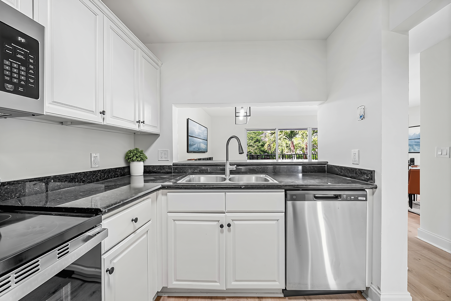 White kitchen cabinets, dark granite countertops, and stainless steel appliances, with a window view to another room.