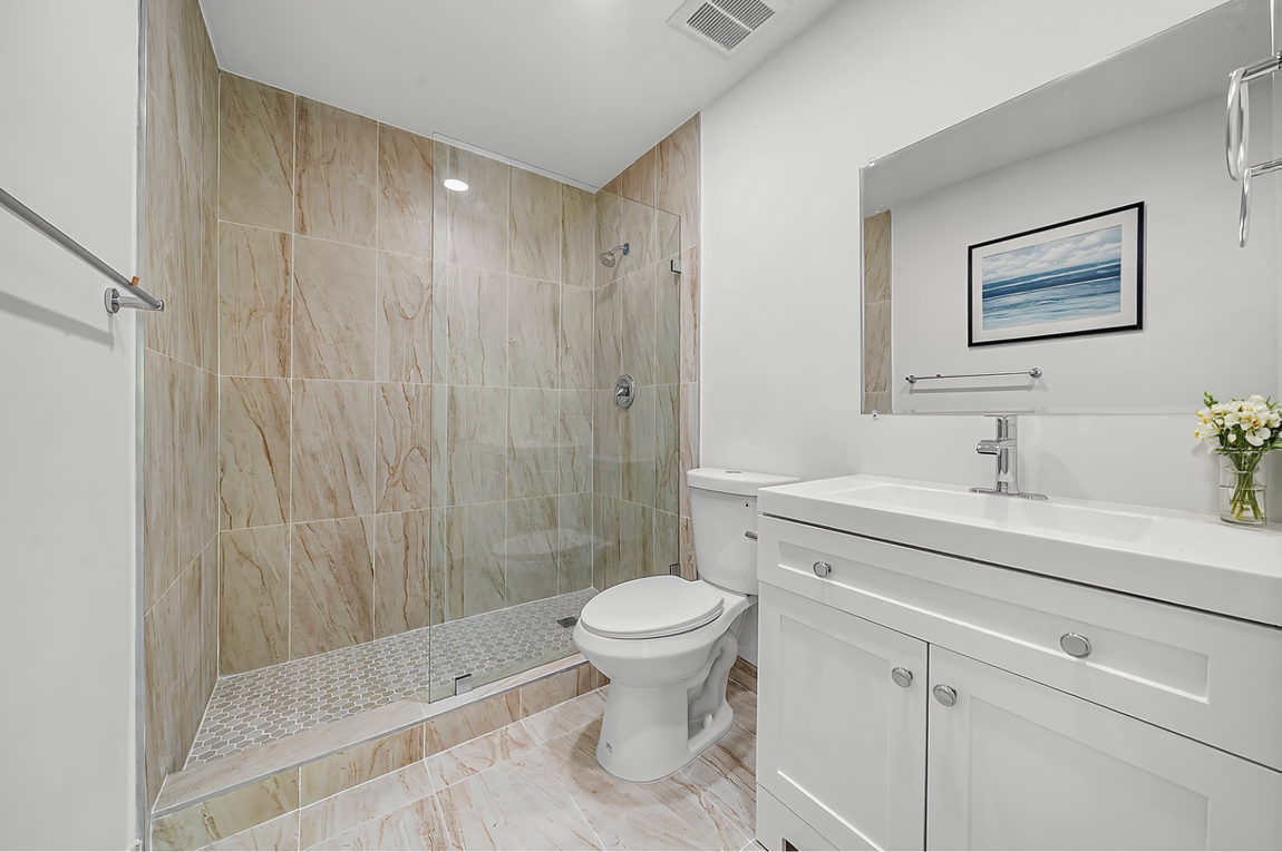 Modern bathroom with a glass-enclosed walk-in shower, light tile walls, white vanity, and a toilet.