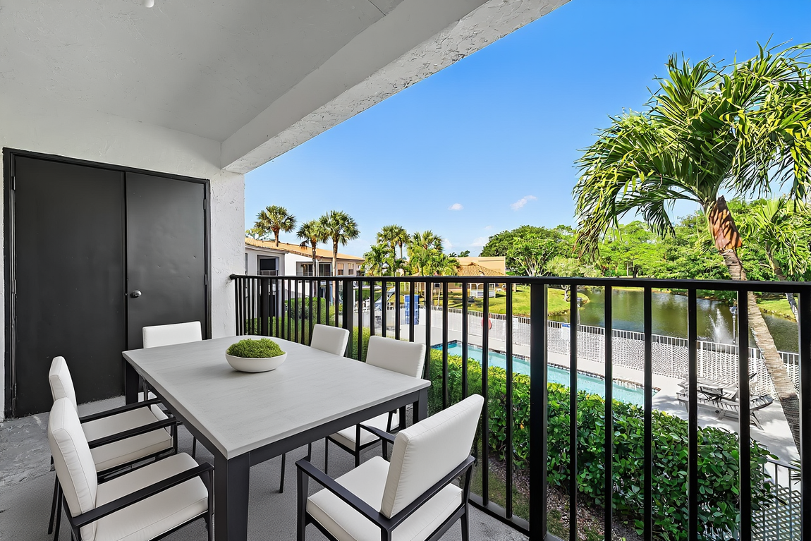 A patio with a dining table and four chairs, overlooking a swimming pool surrounded by palm trees and greenery.