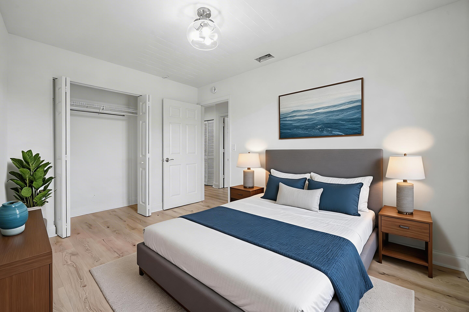 A staged bedroom featuring a grey bed with blue accents, a wooden nightstand with a lamp, and a closet with white doors.
