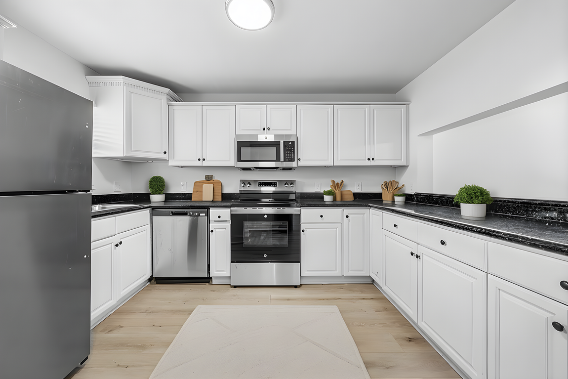 A bright kitchen featuring white cabinets, stainless steel appliances, dark countertops, and light wood-look flooring.