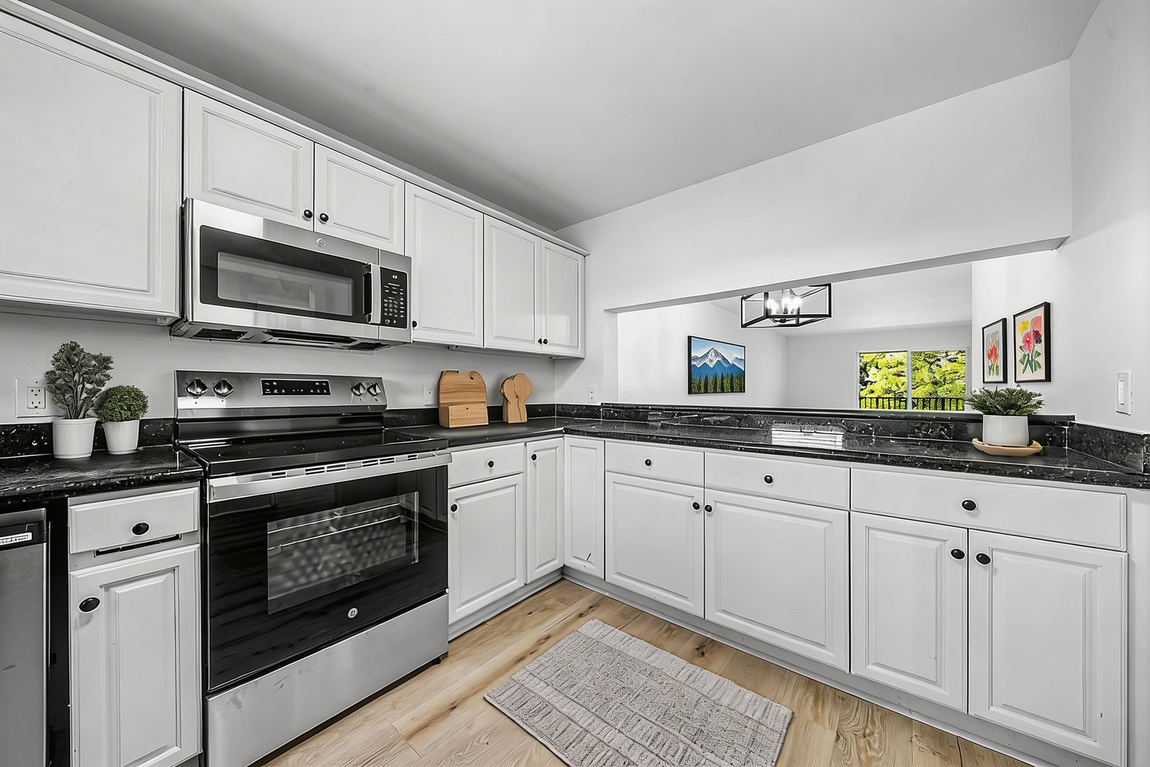 A bright kitchen with white cabinets, dark granite countertops, stainless steel appliances, and wood-style flooring.