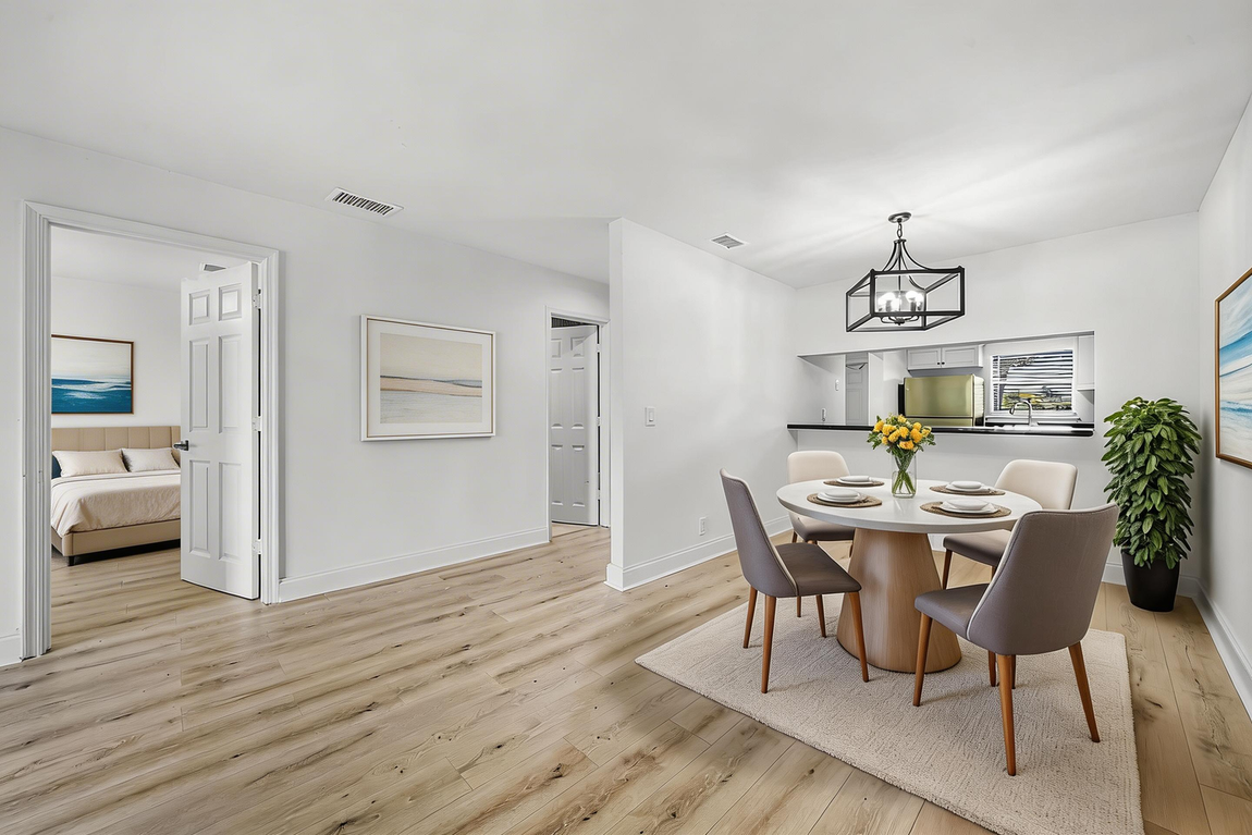 A dining area with a round table and chairs on a rug, adjacent to a hallway leading to a bedroom with light wood floors.