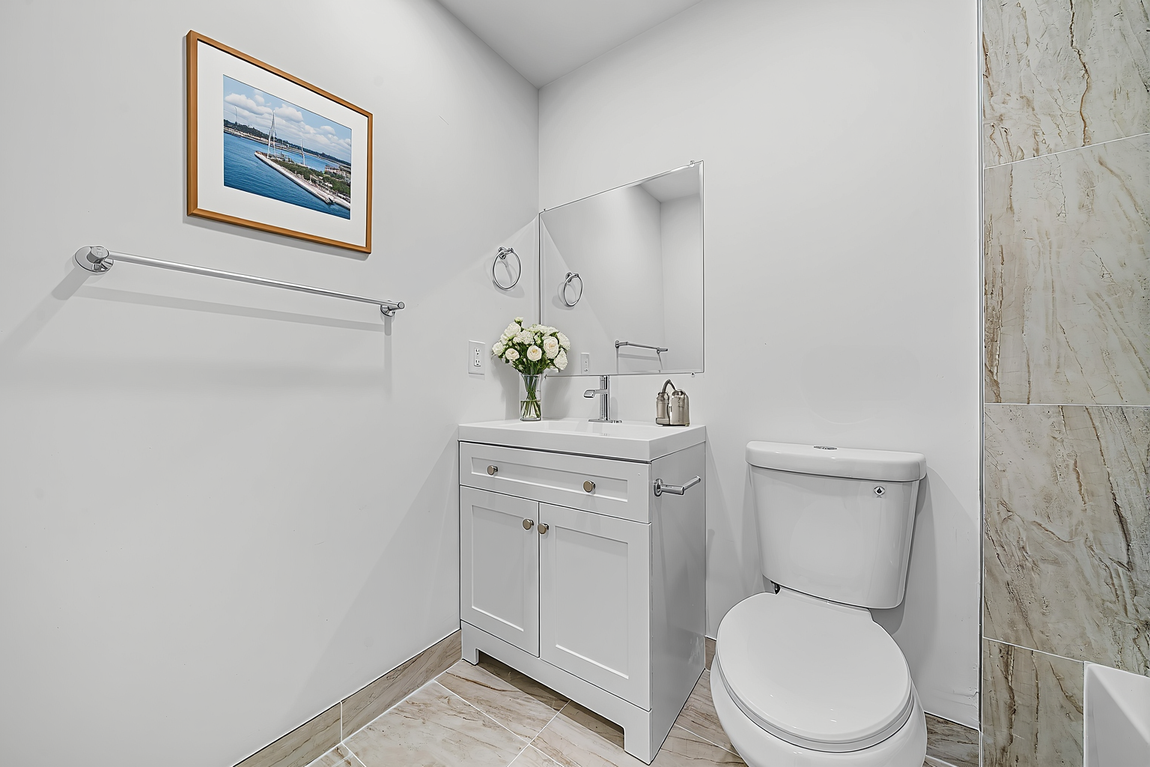 A small, bright bathroom with white walls, a white vanity cabinet, a mirror, a toilet, and framed artwork on the wall.