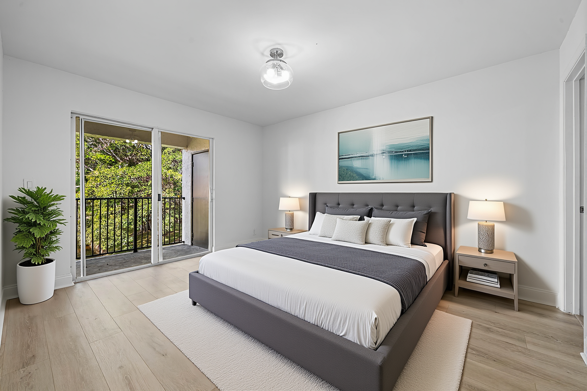 A bright bedroom with a gray tufted bed, white walls, wood floors, a painting above the bed, and a door to a balcony.