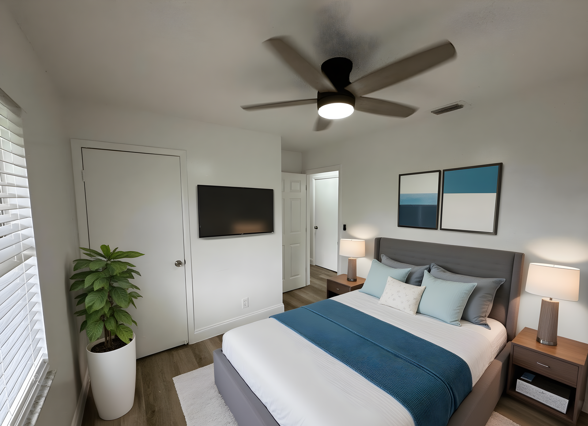 A bedroom with a grey bed, blue accents, a ceiling fan, a potted plant, and a wall-mounted TV.
