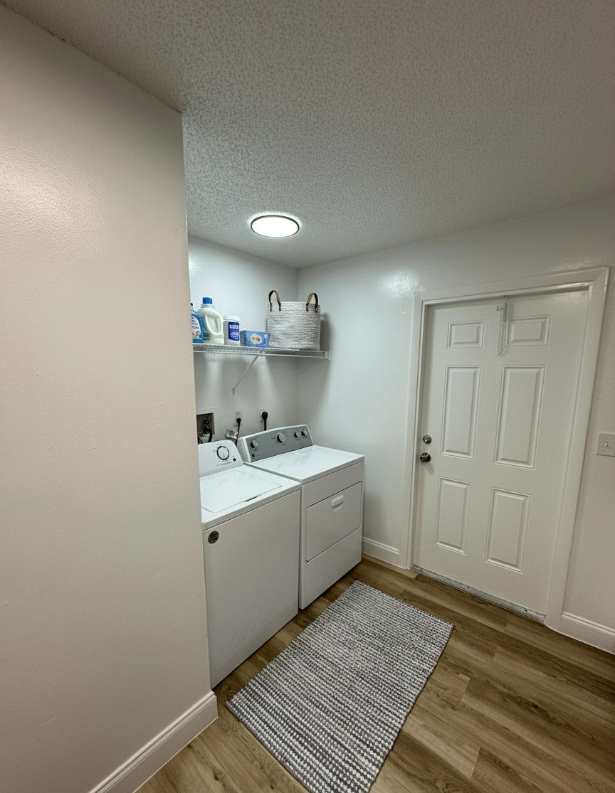 A white laundry room featuring a washer and dryer, a storage shelf with supplies, and a door on a light wood floor.