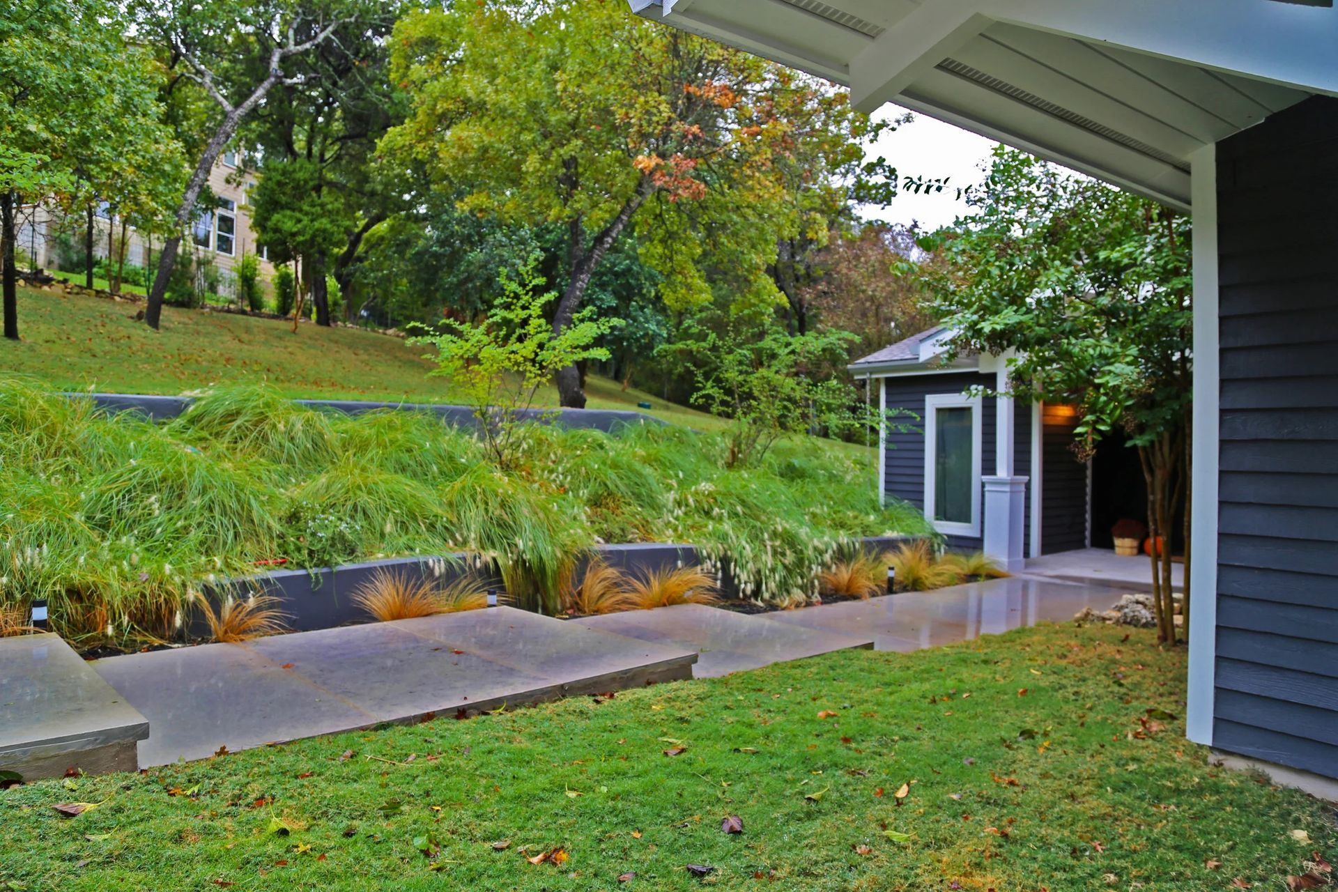 Lawn, tiered planters with tall grasses, and a dark-blue building under a white overhang. Trees in background.