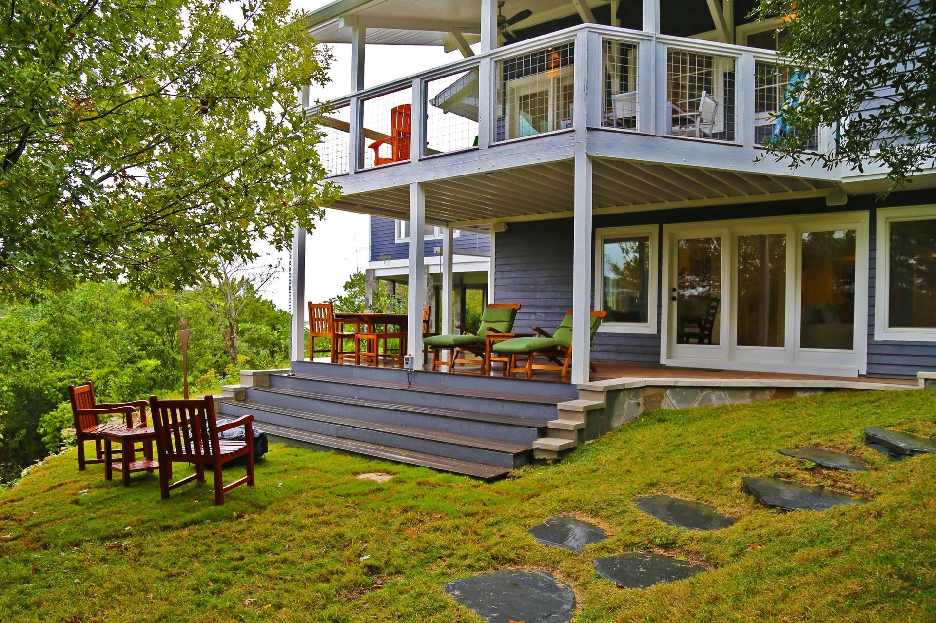 Two-story blue house with porch and deck overlooking a grassy yard with outdoor seating.