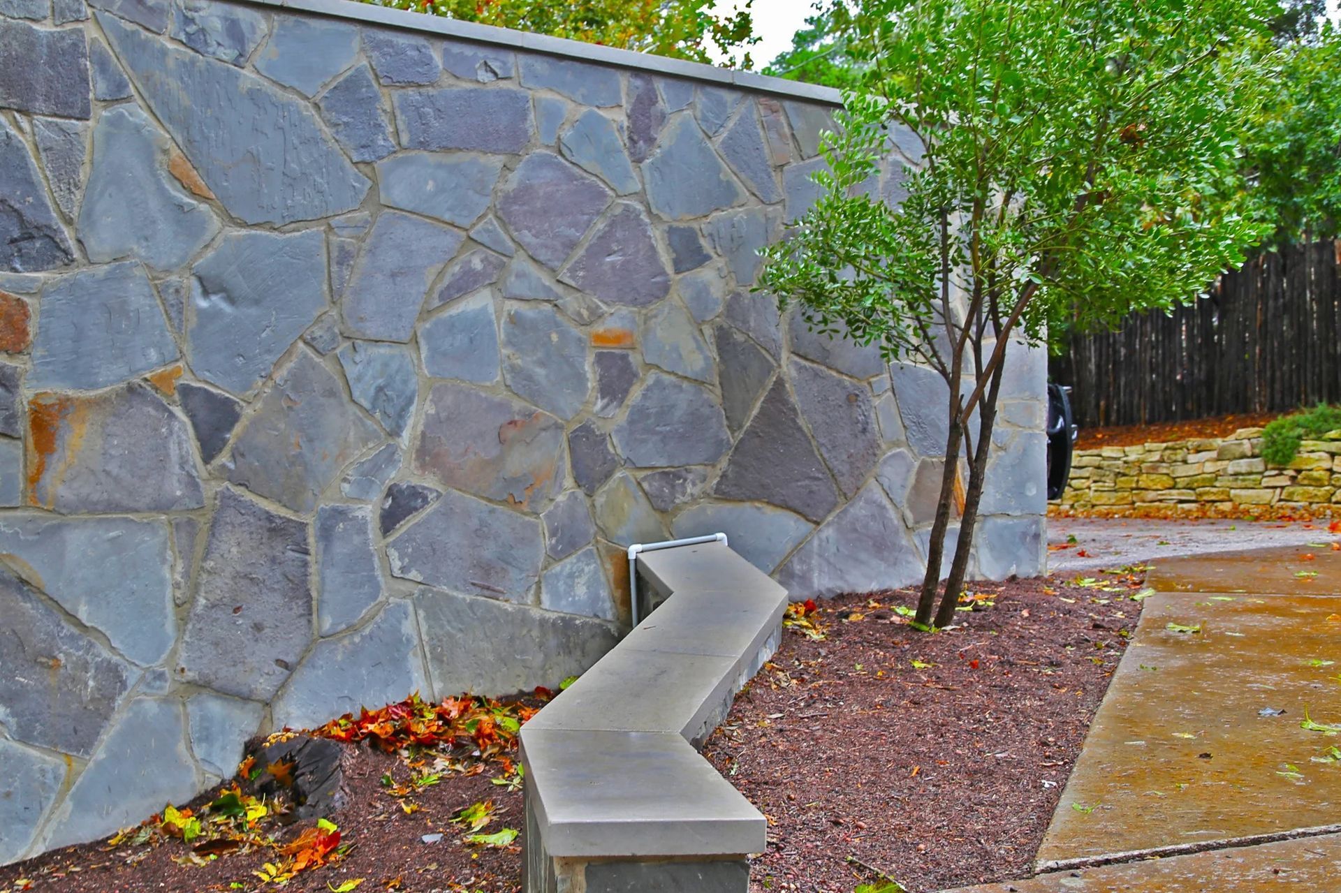 Stone wall with zigzag concrete bench, tree, and walkway.