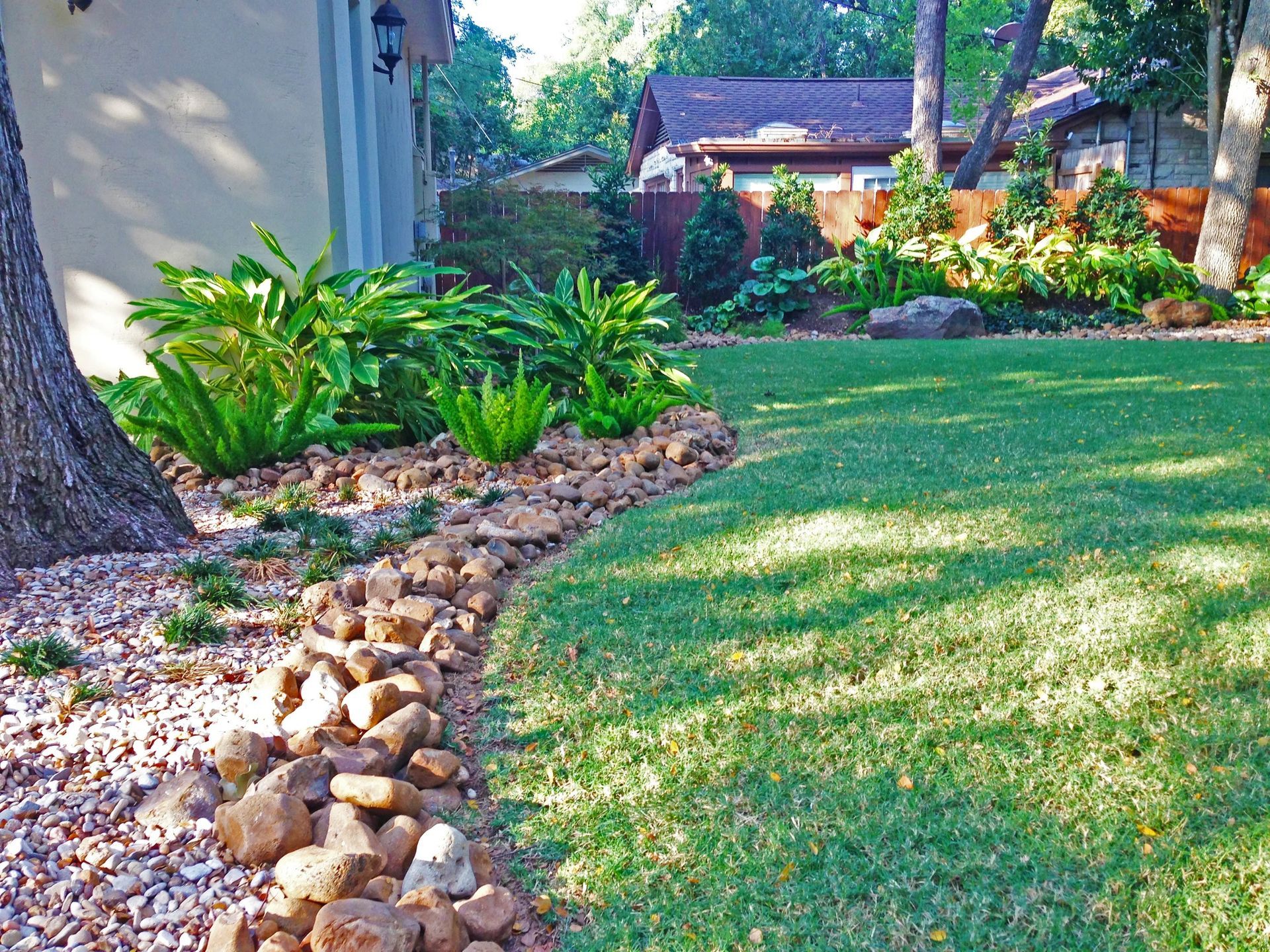 Rock border separates a lawn and garden with lush greenery and a tan house.