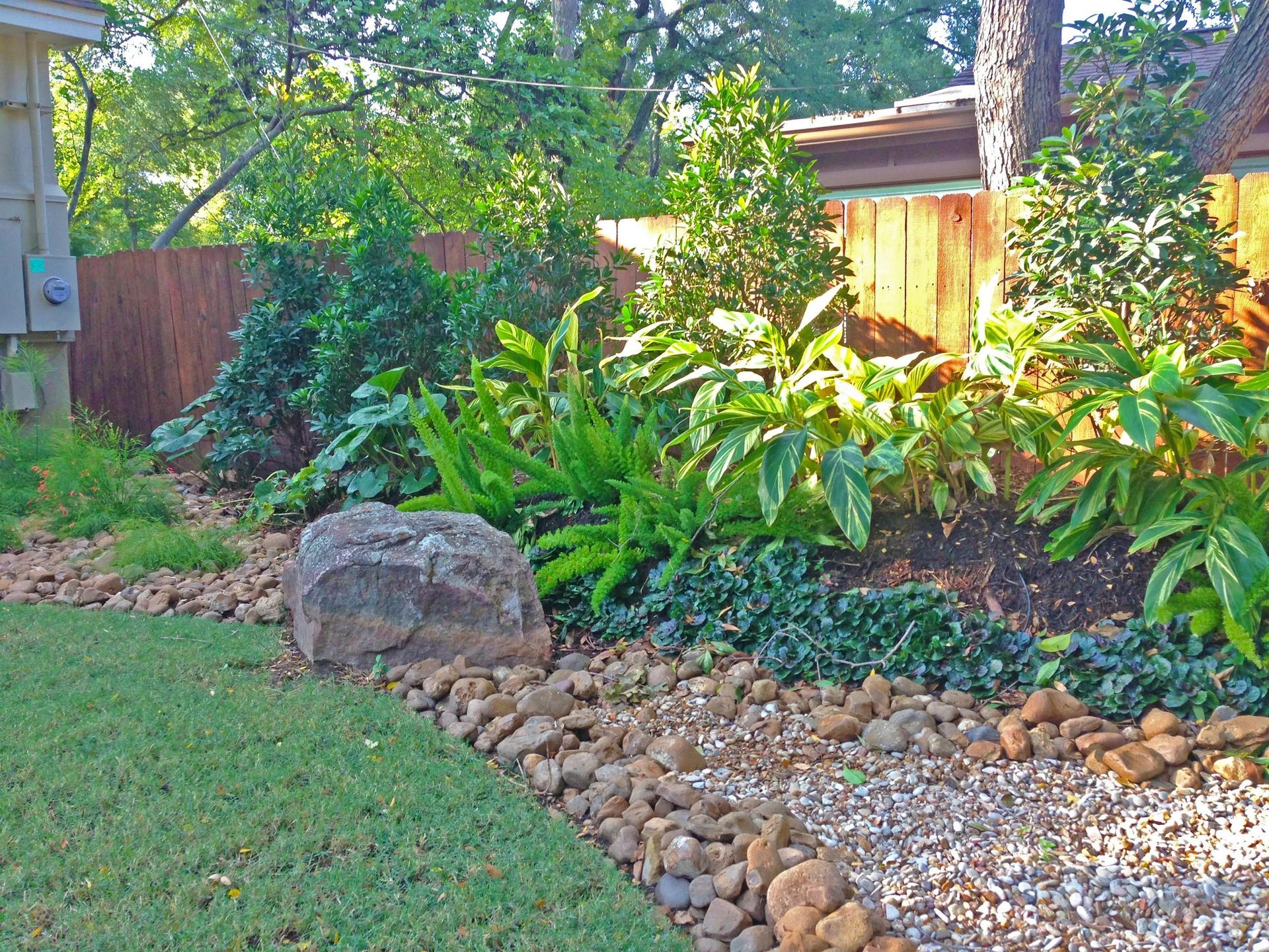 Lush garden bed with large rock border, containing green plants and brown gravel. Fence in the background.