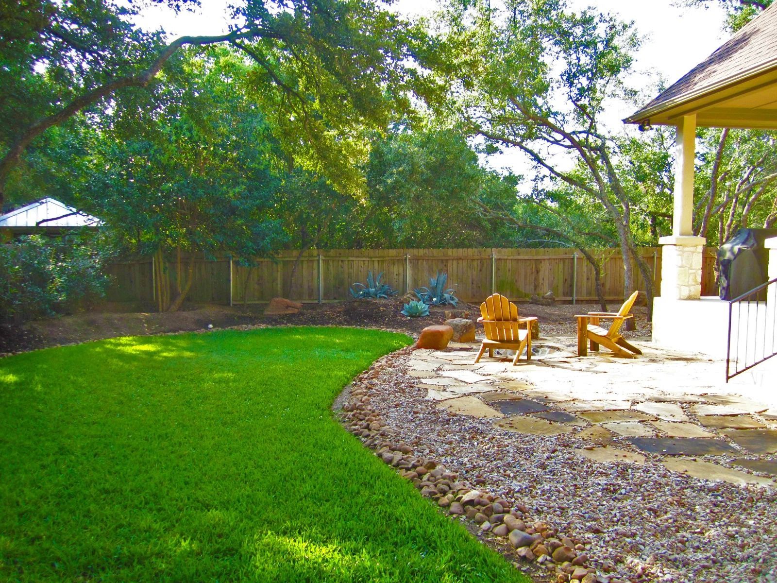 Green lawn and stone patio with wooden chairs, next to a rock bed and wooden fence, under trees.
