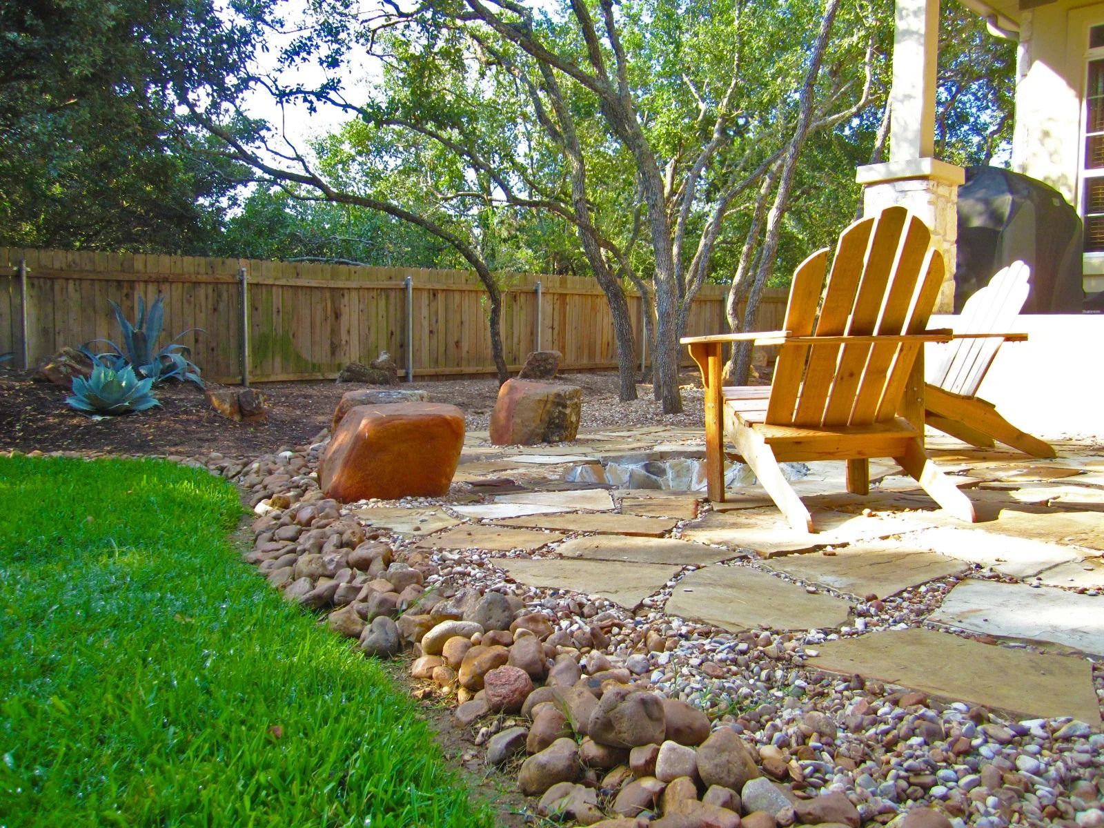 Wooden Adirondack chair on a stone patio in a backyard with a rock border and wooden fence.