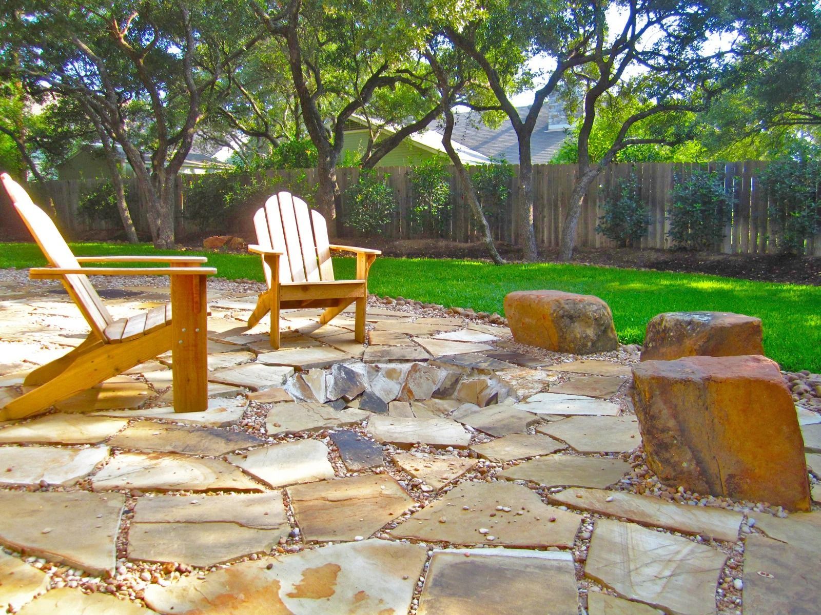 Two wooden Adirondack chairs face a stone patio with a fire pit, in a backyard setting with trees and a wooden fence.