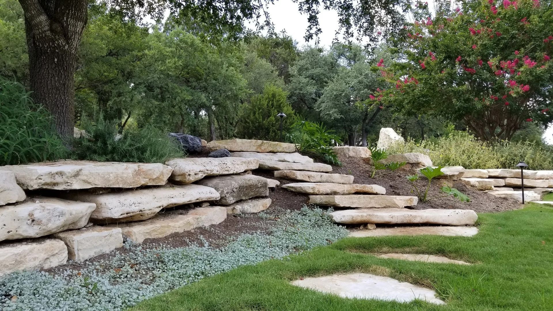 Stone steps in a landscaped yard. Green grass and plants with trees in the background.