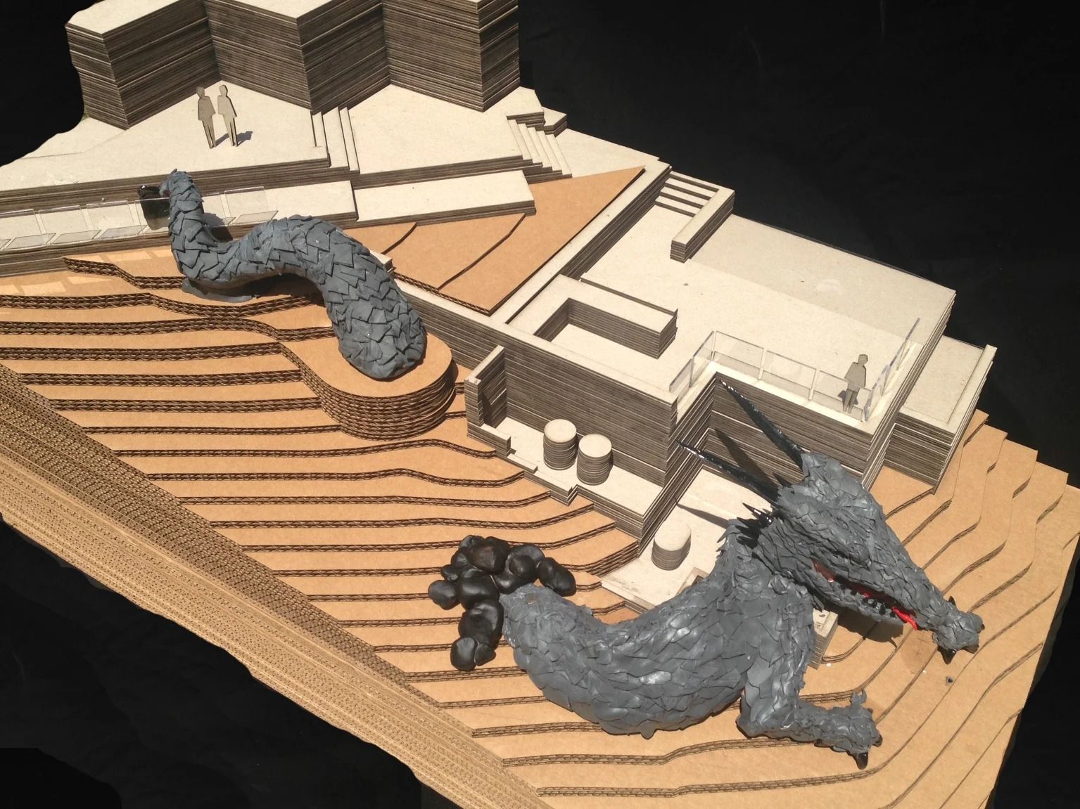 Cardboard model of a building with a gray, dragon-like sculpture and a tail winding up a hill.