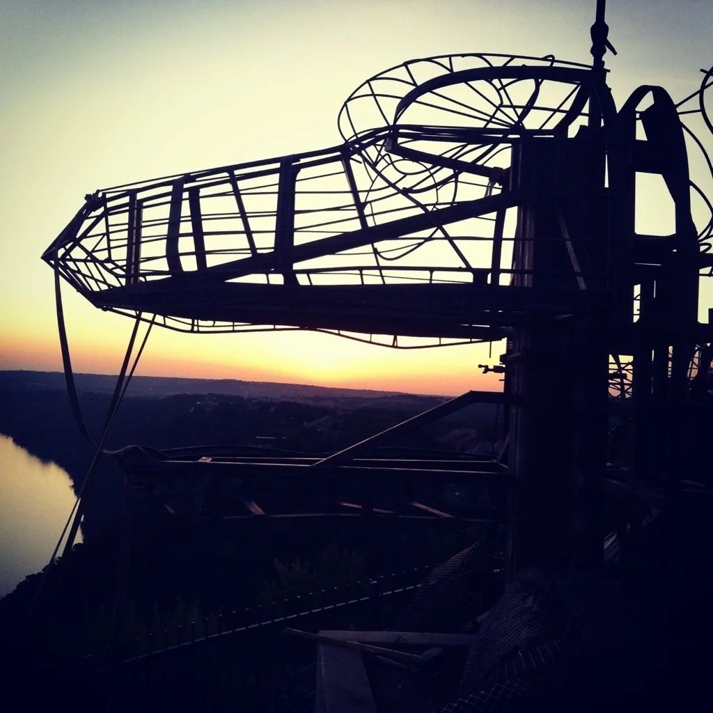 Silhouette of a metal sculpture resembling a dinosaur head at sunset, overlooking a river.