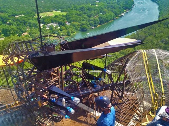 Construction worker near large, metal sculpture with black wings, overlooking a river.
