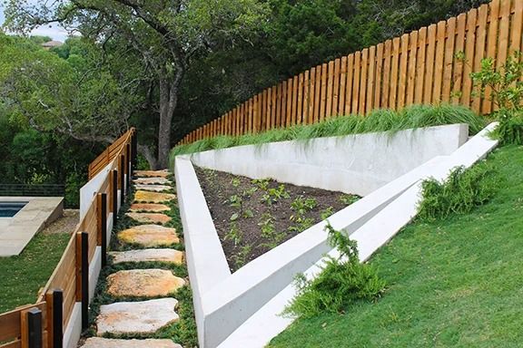 Stone steps lead up a grassy slope, past a concrete planter to a wooden fence.