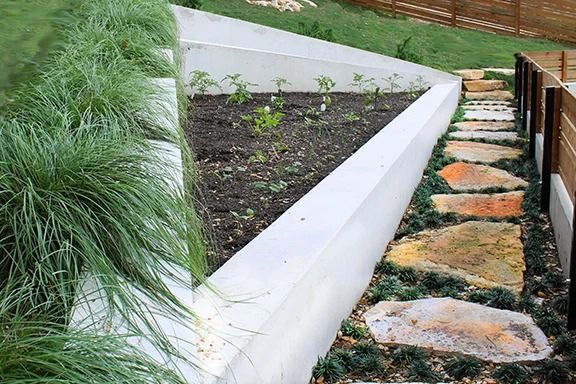 Raised concrete garden bed with stone path, green grass, and small plants.