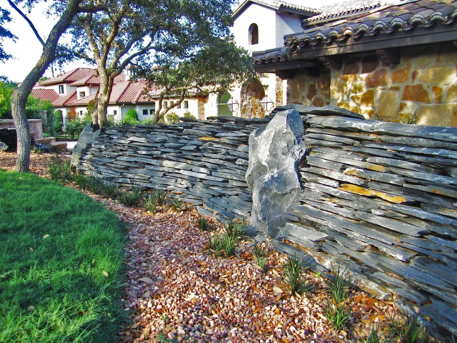 Stone wall in front of a building with red-tiled roof. Green grass and a path with fallen leaves.