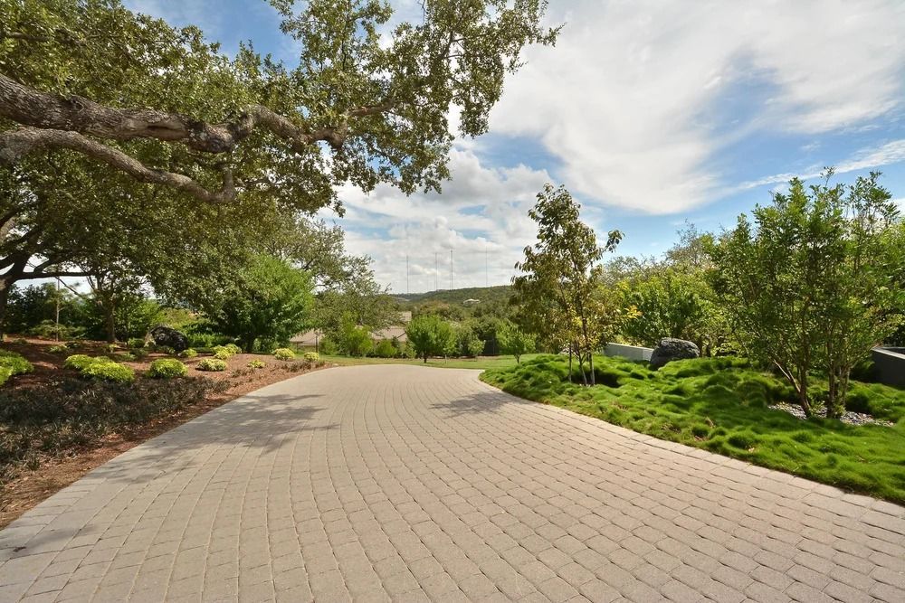 Brick driveway winds through lush greenery, trees, and blue sky with clouds.