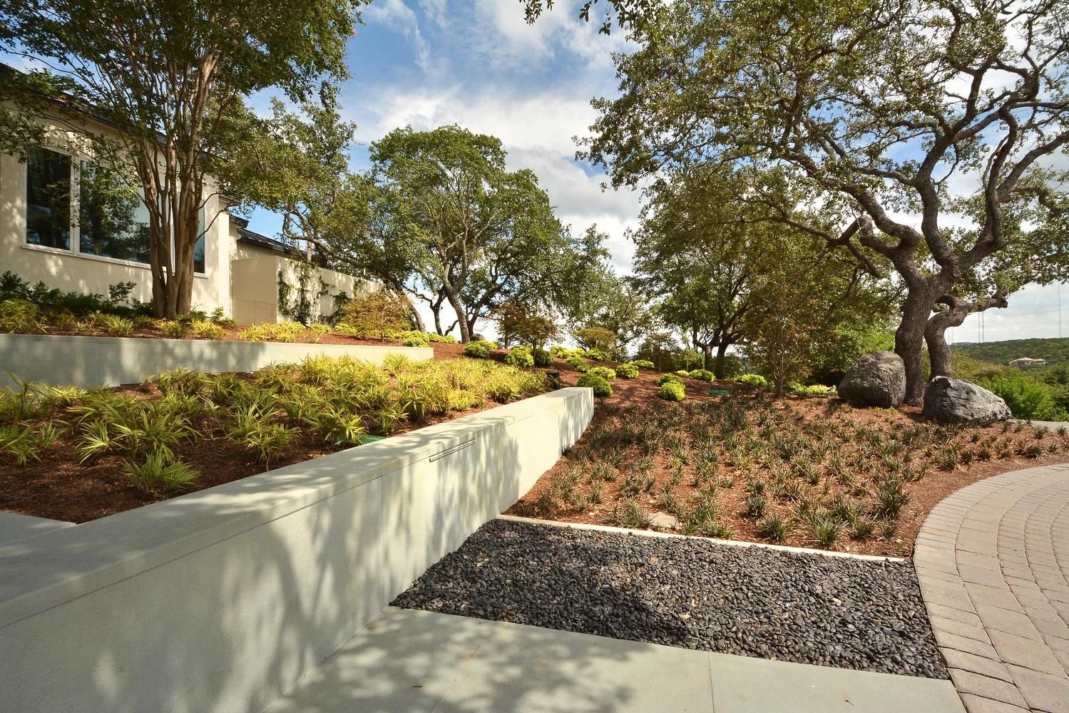 Landscaped yard with a house, retaining walls, trees, plants, and a circular driveway. Sunny day.