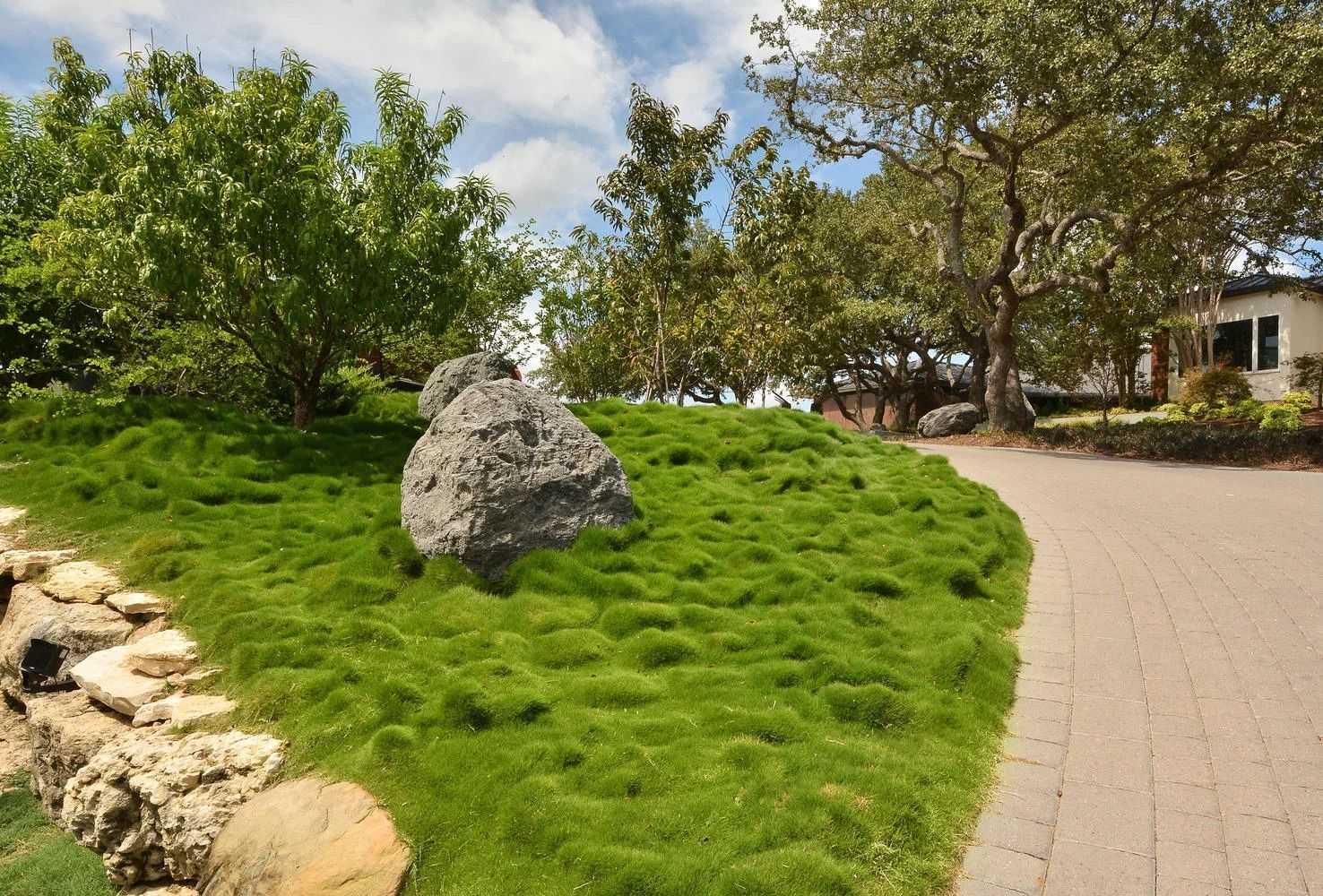 Lush green grass hill with a large rock, trees, and a brick pathway under a partly cloudy sky.