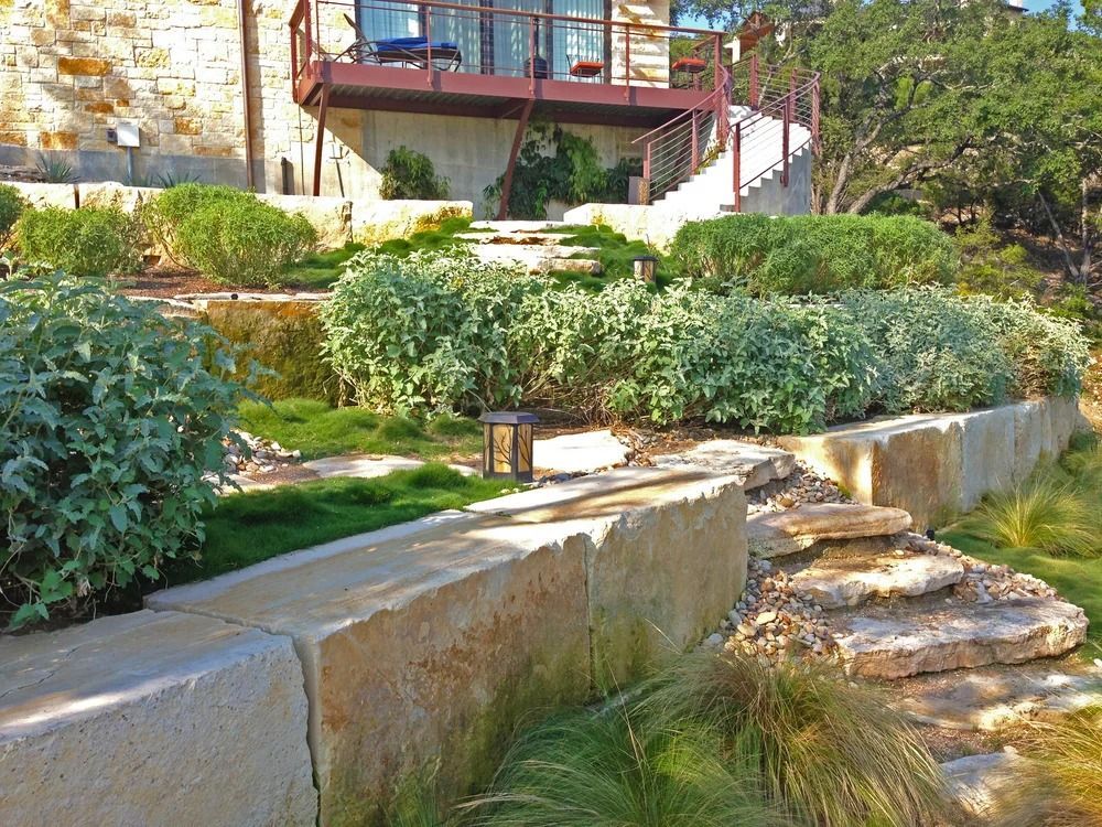 Stone retaining walls and steps in a tiered landscape leading up to a house with a balcony.