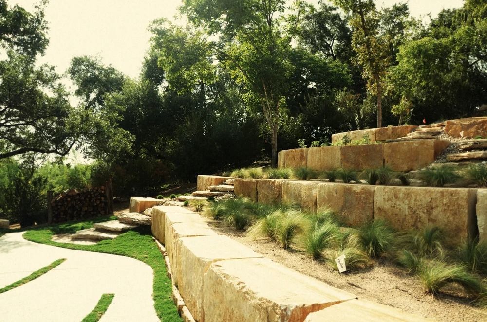 Stone terraced garden with grass and shrubs, surrounded by trees.