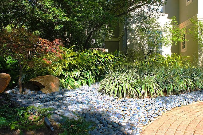 Garden bed with rocks and lush plants, next to a brick path and building.