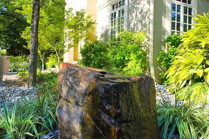 Water fountain in a lush garden setting. A light-colored building in the background.